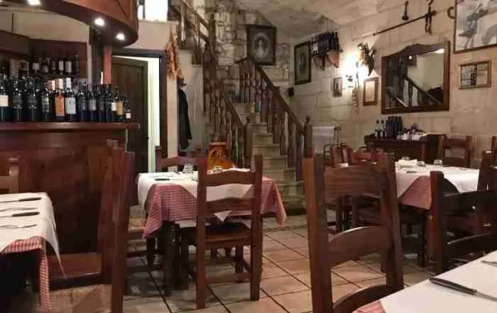 Vista d'insieme della saletta: muri in pietra, scala in legno, tavoli apparecchiati con tovaglie bianche e a quadretti rossi